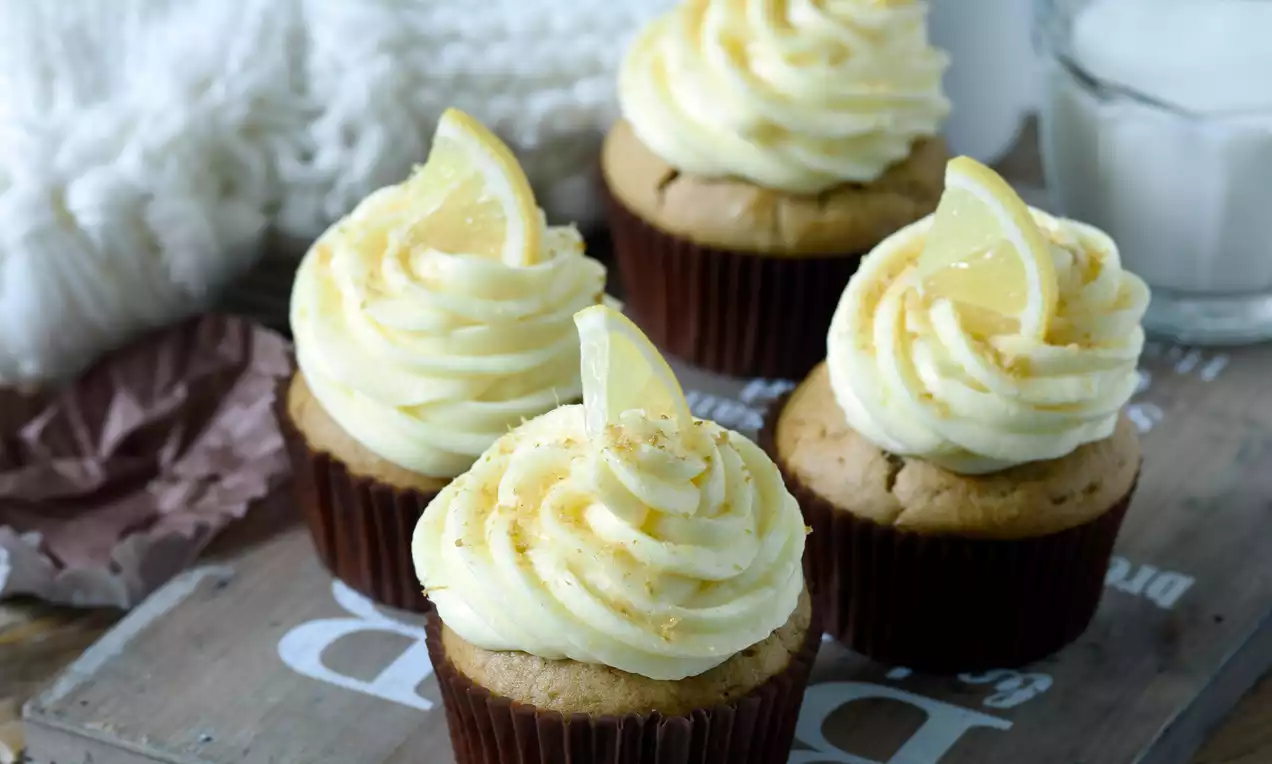Veganské muffiny s krémom svetlohnedej farby v tmavých košíčkoch, ozdobené nadýchaným bledožltým krémom a plátkami citróna na drevenom podnose