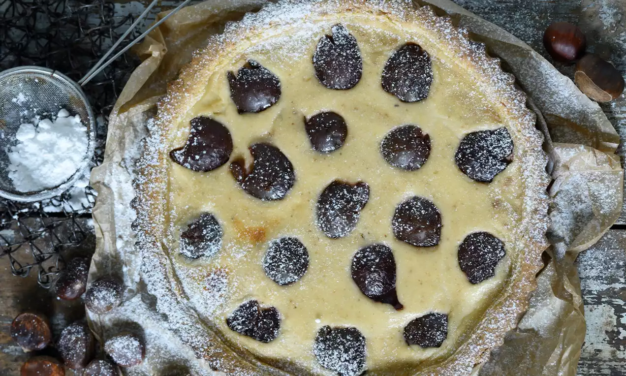 Tart s jemne zlatistým okrajom, hladkou plnkou krémovej farby, zdobený polovičkami tmavých sliviek a práškovým cukrom na papieri na pečenie