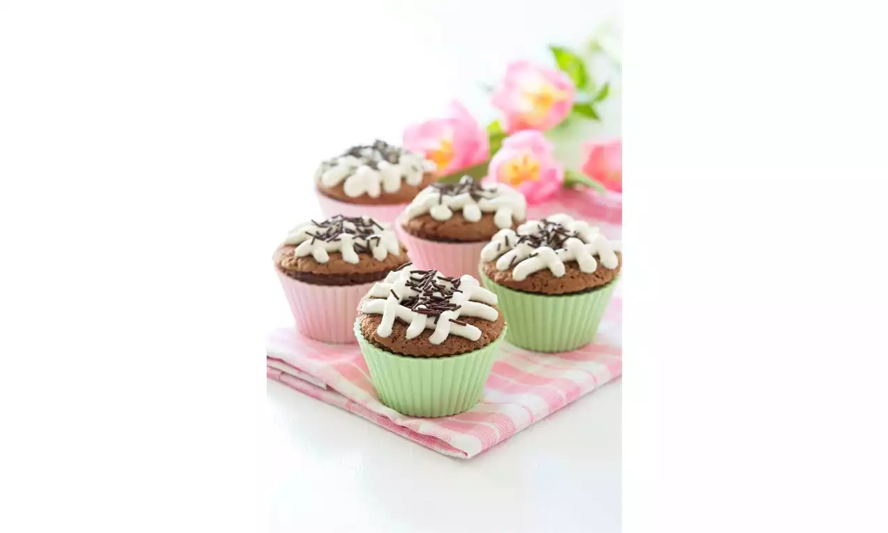 Čokoládové cupcakes v farebných košíčkoch s bielou polevou a hoblinkami čokolády na jemnej ružovo-bielej utierke vedľa ružových kvetov