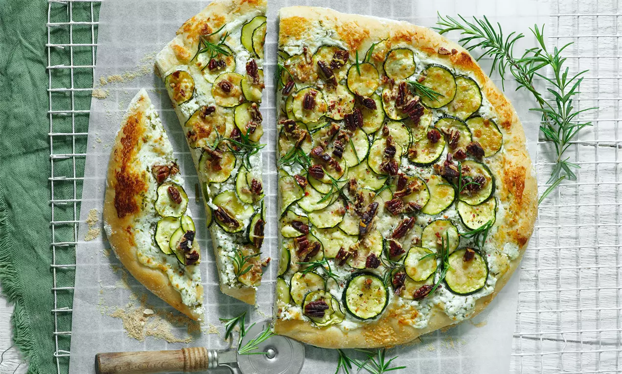 Cuketová pizza so zlatistým okrajom, zelenými plátkami cukety, hnedými kúskami orechov, bylinkami a krémovou bielou základňou na bielom pergamene