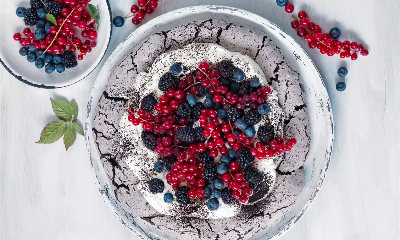 Snehovo biela Pavlova s prasknutým kakaovým povrchom, korunovaná lesklou červenou ríbezľou, černicami a čučoriedkami na bledom tanieri