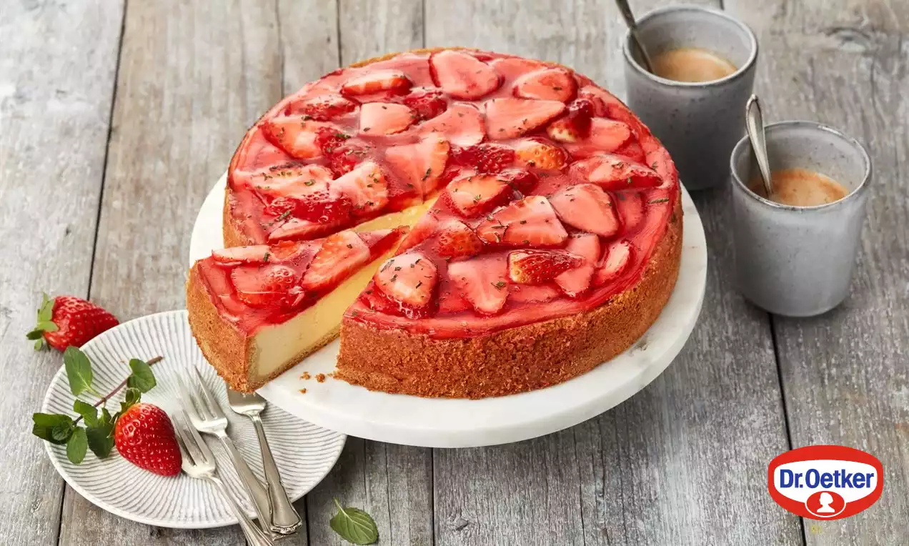 Cheesecake s hladkým krémovým stredom na zlatohnedom korpuse, pokrytý lesklými plátkami čerstvých jahôd a červeným želé, servírovaný na bielom tanieri