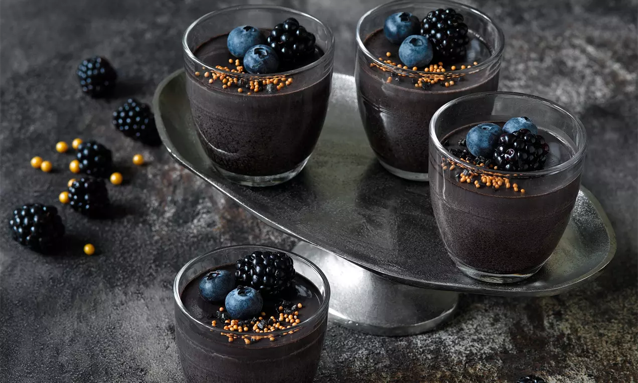 Lesklá čierna panna cotta v tvare hladkého zaobleného valca na tmavom rustikálnom podklade, ozdobená černicami a zlatými cukrovými perličkami