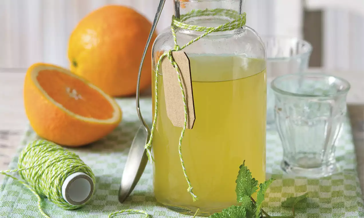 Medovkovo-zelený sirup v sklenenej fľaši s papierovým štítkom, ozdobený zeleným špagátom, vedľa rozpoleného pomaranča a lístkov medovky