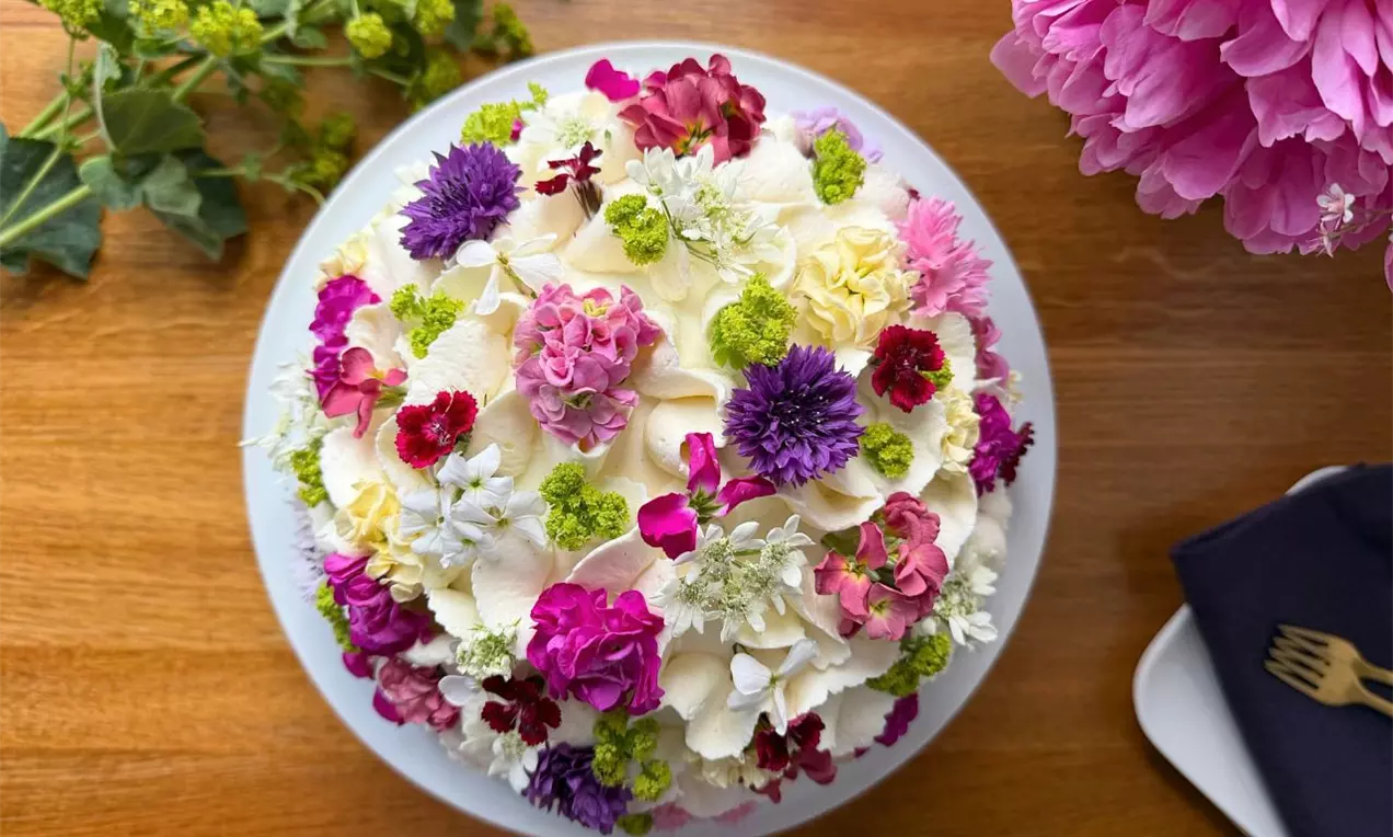 Okrúhla torta na bielom tanieri zdobená pestrými kvetmi v odtieňoch ružovej, fialovej, bielej a zelenej, ležiacej na drevenom stole s kvetinovou dekoráciou