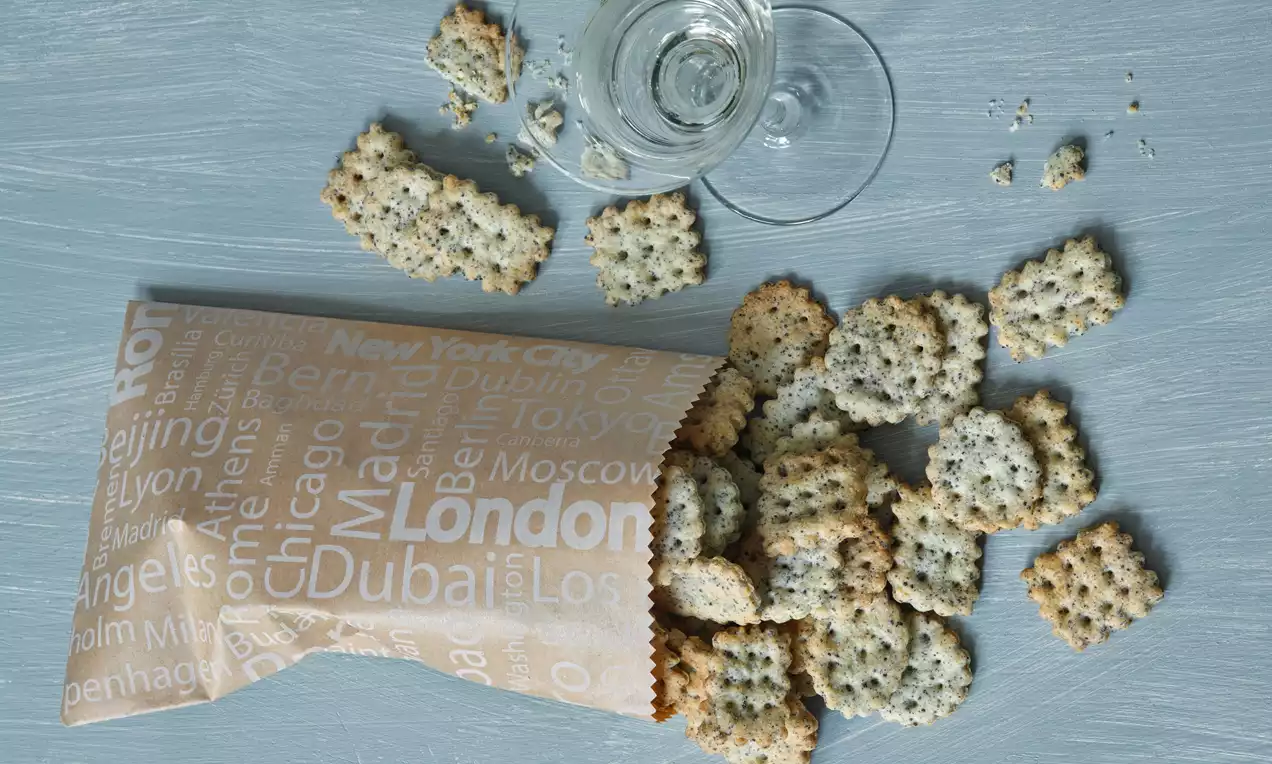 Slané krekry štvorcového tvaru so zlatistými okrajmi, posypané čiernym makom, rozložené vedľa pohára vína na modrom drevenom povrchu