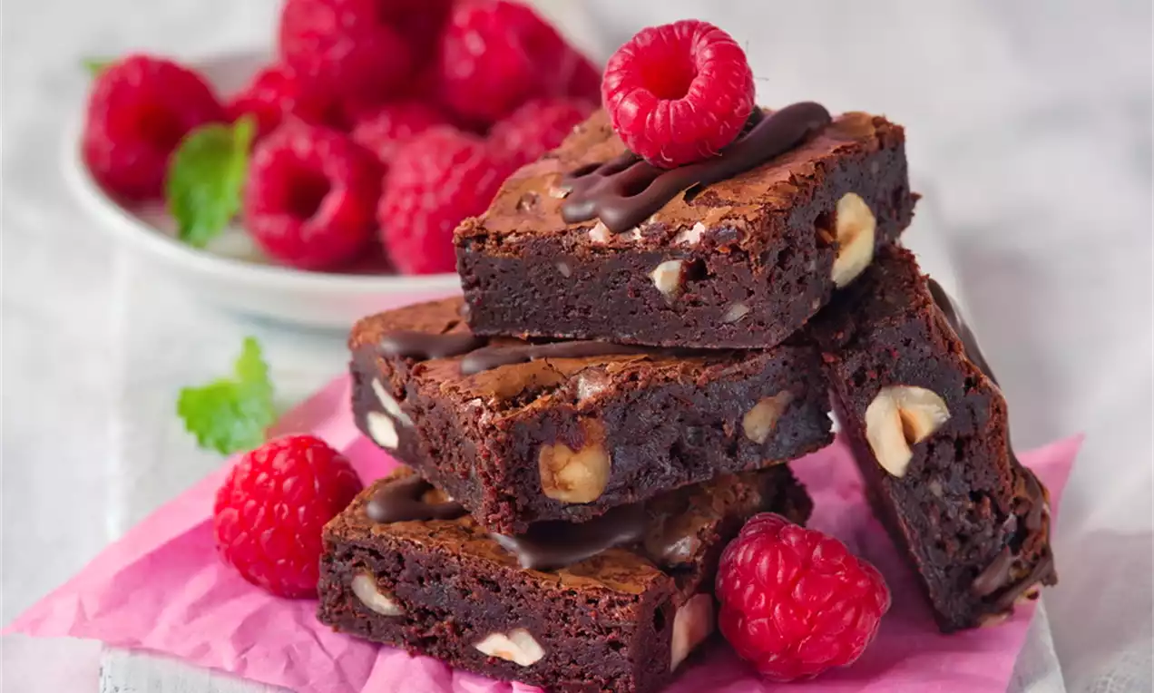 Kúsky tmavohnedých brownies s orechmi, uložené na sebe na ružovom papieri, ozdobené čerstvými malinami a jemne zozadu osvetlené
