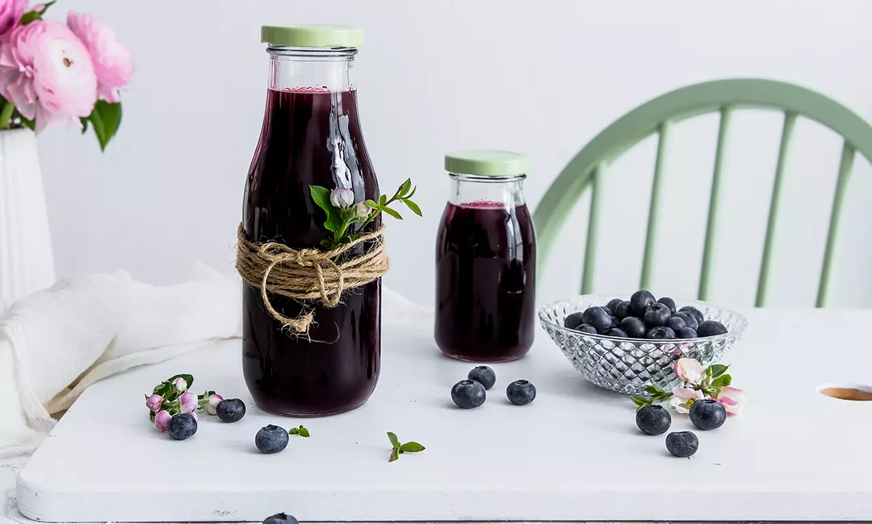 Tmavofialový čučoriedkový sirup v štíhlych sklenených fľašiach s zeleným uzáverom, ozdobený šnúrkou a vetvičkou, obklopený čerstvými čučoriedkami