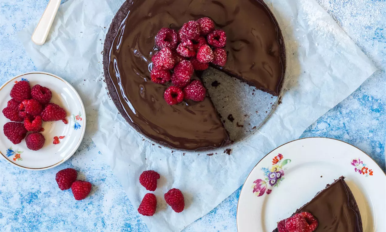 Čokoládová torta tmavohnedej farby s lesklým povrchom, navrchu zoskupené čerstvé červené maliny, servírovaná na bielom tanieri s kvetinovým vzorom