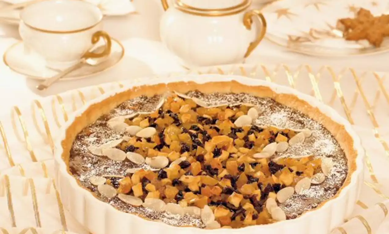 Adventný quiche s hnedou náplňou posypanou kockami sušeného ovocia, plátkami mandlí a práškovým cukrom v bielej porcelánovej forme