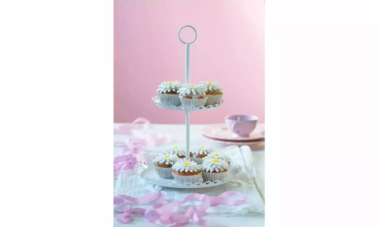 Citrónové muffiny s nadýchanou bielou polevou a ozdobou v tvare sedmokrások, naaranžované na dvojposchodovom bielom stojane so stužkou a šálkou v pozadí