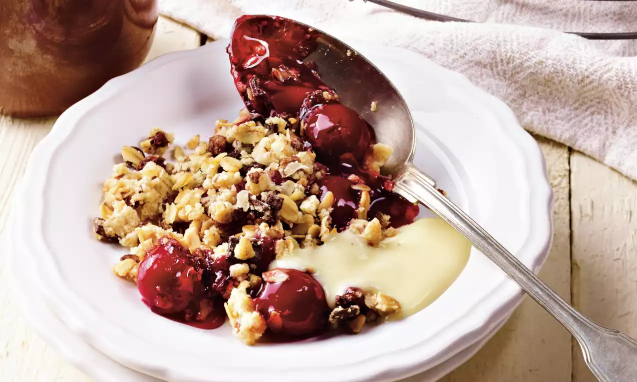 Teplý višňový crumble s červenými šťavnatými višňami, zlatistou mrveničkou a krémovou vanilkovou omáčkou na bielom porcelánovom tanieri