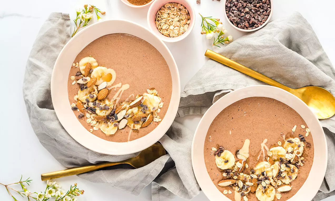 Banánová smoothie miska svetlohnedej farby, ozdobená plátkami banánu, čokoládovými kúskami, orieškami a drizzlom krémovej polevy na bledom pozadí