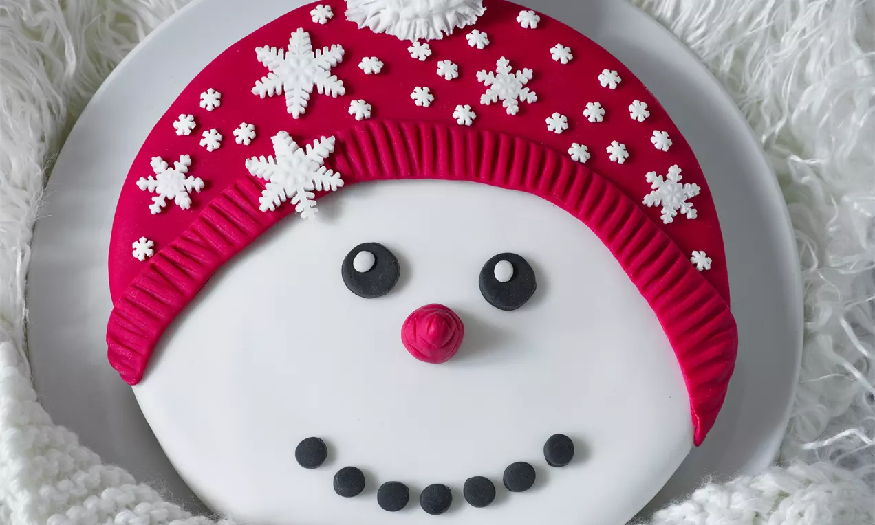 Biela torta v tvare snehuliaka so zvýraznenou červenou čiapkou, ozdobená malými snehovými vločkami, čiernymi očami a úsmevom z bodiek