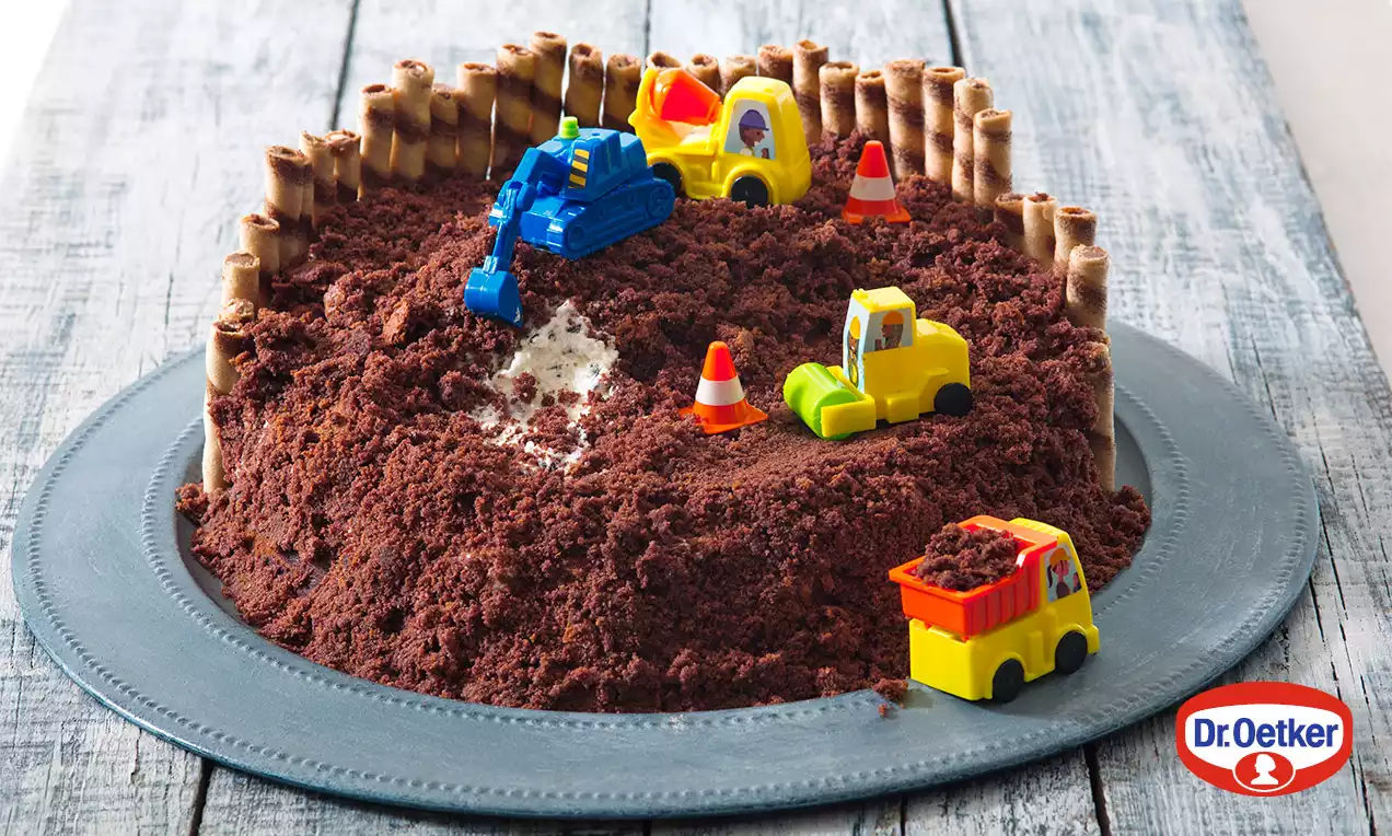 Čokoládová kopa roztrúsená na tmavom oblom torte obloženom trubicovými tyčinkami, s detskými strojmi a kužeľmi evokujúca stavenisko