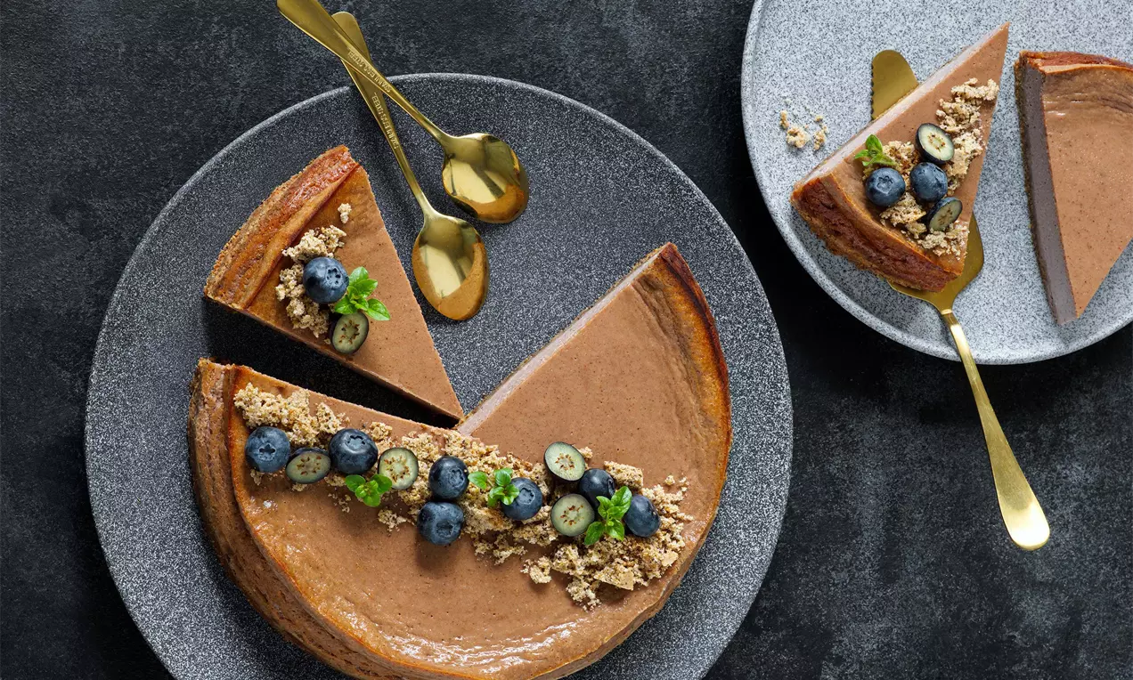 Makový cheesecake s hladkým kakaovým krémom na tmavom korpuse, ozdobený čučoriedkami, drobenkou a lístkami mäty, na šedom tanieri s lyžičkami