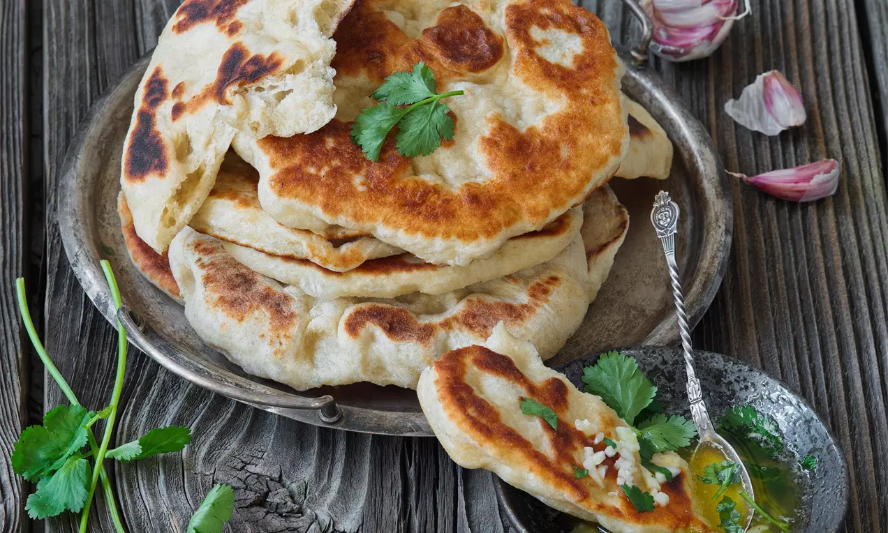 Zlatohnedé indické placky naan s ľahko opečenými povrchmi, položené na kovovom tanieri, zdobené lístkami koriandra vedľa misky s cesnakovým maslom
