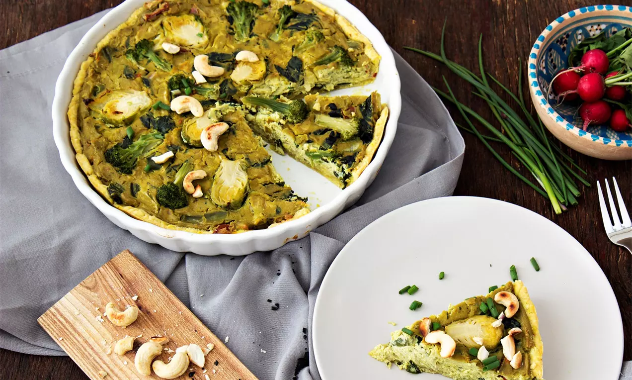 Vegánsky zeleninový quiche v zlatohnedom ceste, s plnkou zo zelenej zeleniny a kešu orieškami, podávaný na bielom tanieri so zelenou pažitkou