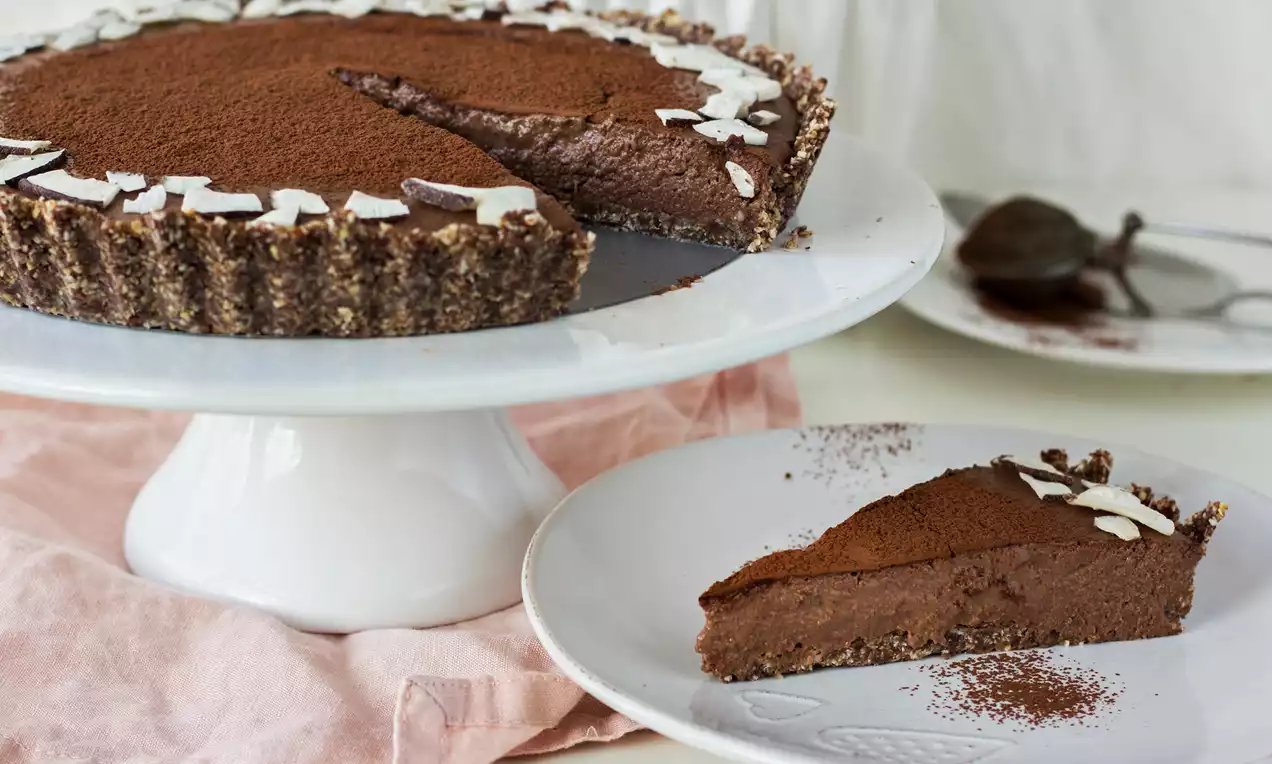 Čokoládovo-kokosový tart s tmavohnedou čokoládovou plnkou na orechovo-kokosovom korpuse, posypaný kakaovým práškom a plátkami kokosu na bielom tanieri.