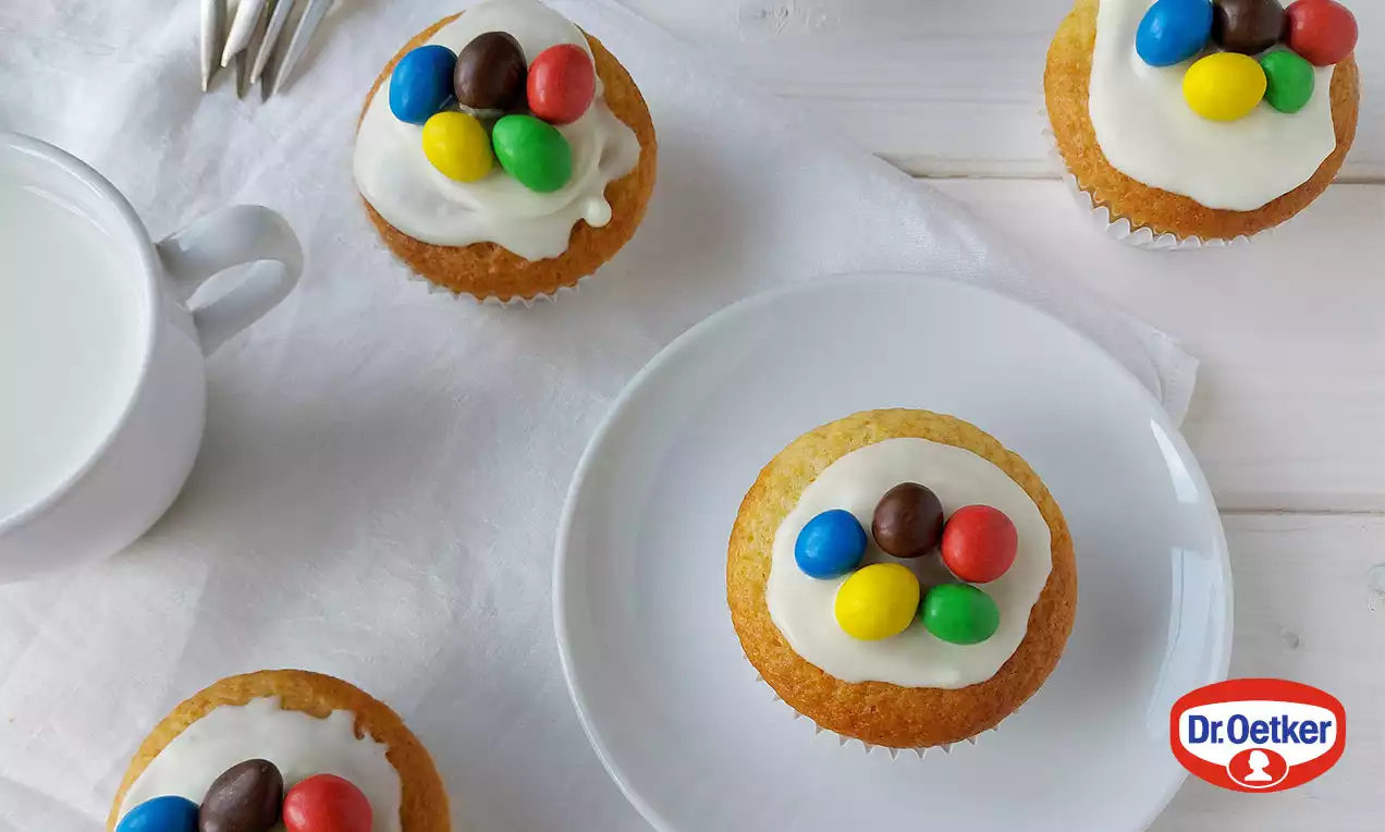 Olympijské muffiny zlatohnedé a nadýchané, s vrstvou bielej polevy ozdobenej pestrofarebnými lentilkami na bielom tanieri s bielym obrúskom