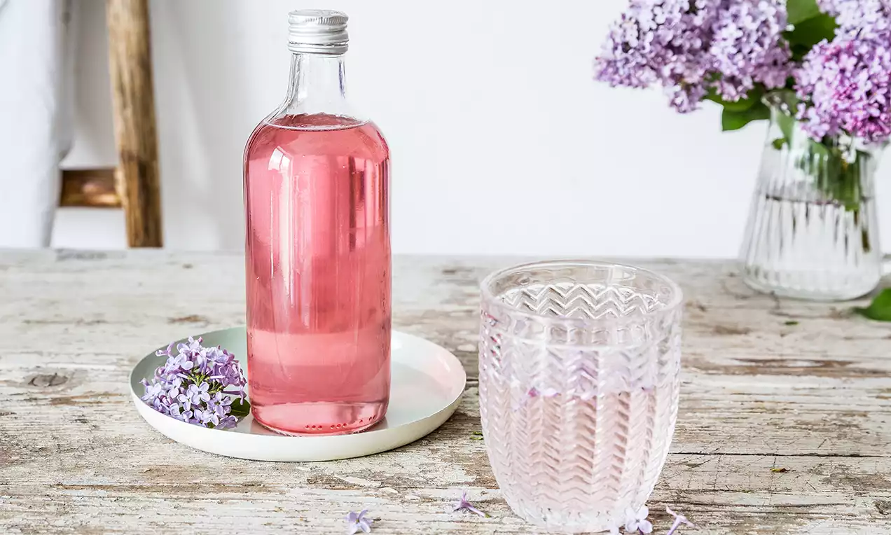 Priezračný ružový sirup v sklenenej fľaši s kovovým uzáverom, vedľa pohára s jemným vzorom, na drevenom stole s dekoráciou fialových kvetov