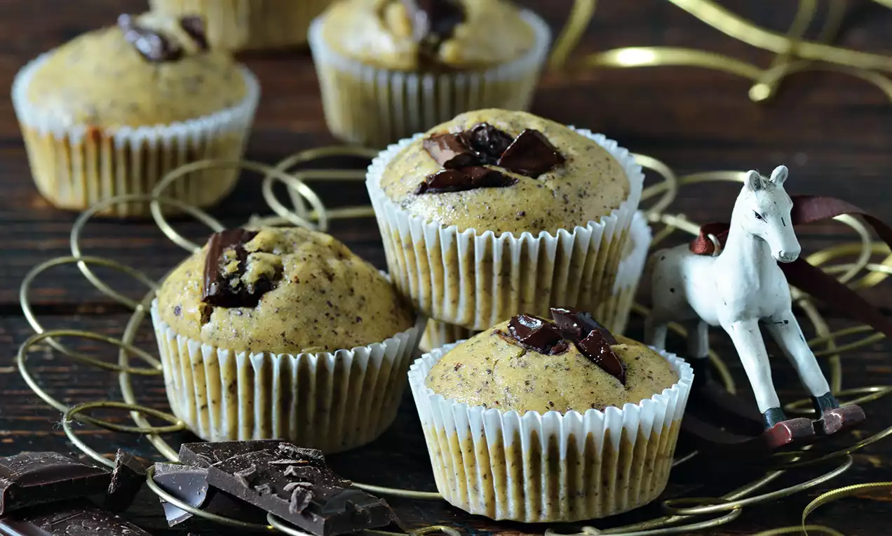 Banánové muffiny v svetlých papierových košíčkoch, posypané kúskami tmavej čokolády, usporiadané na ozdobnom kovovom podnose s hračkou koníka.