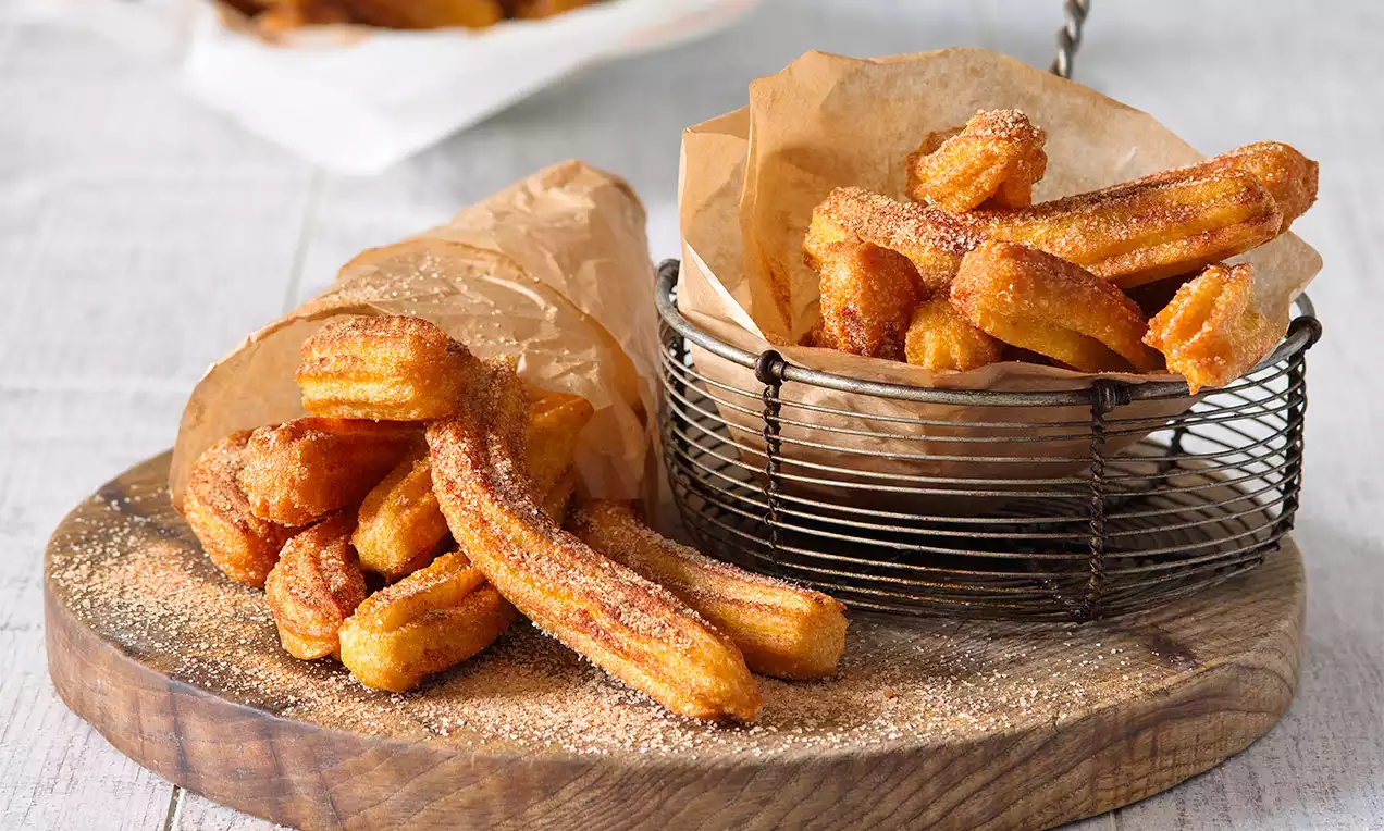 Zlatohnedé churros s chrumkavým povrchom jemne posypané škoricovým cukrom, usporiadané v kovovom košíku a papierovom kornútku na drevenej doske