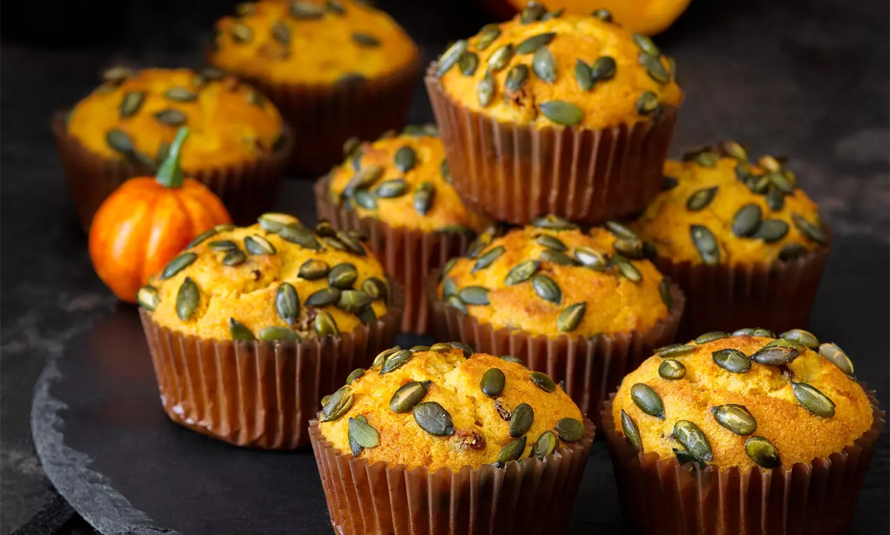 Dýňové muffiny vohnedo-oranžovej farbe, posypané zelenými tekvicovými semiačkami, naaranžované na čiernej podložke s dekoráciou malej tekvice