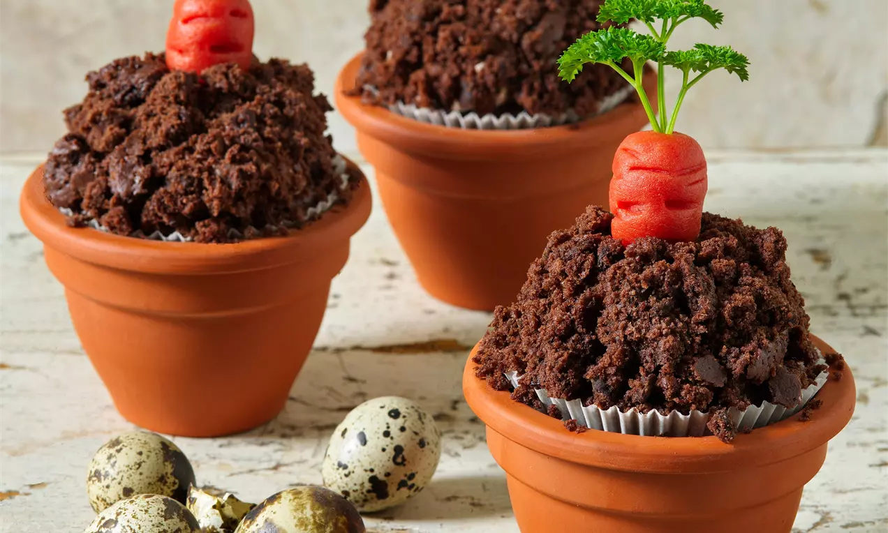 Čokoládové muffiny s tmavohnedou drobivou textúrou, servírované v terakotových kvetináčoch, s marcipánovou oranžovou mrkvou a zelenou vňaťou
