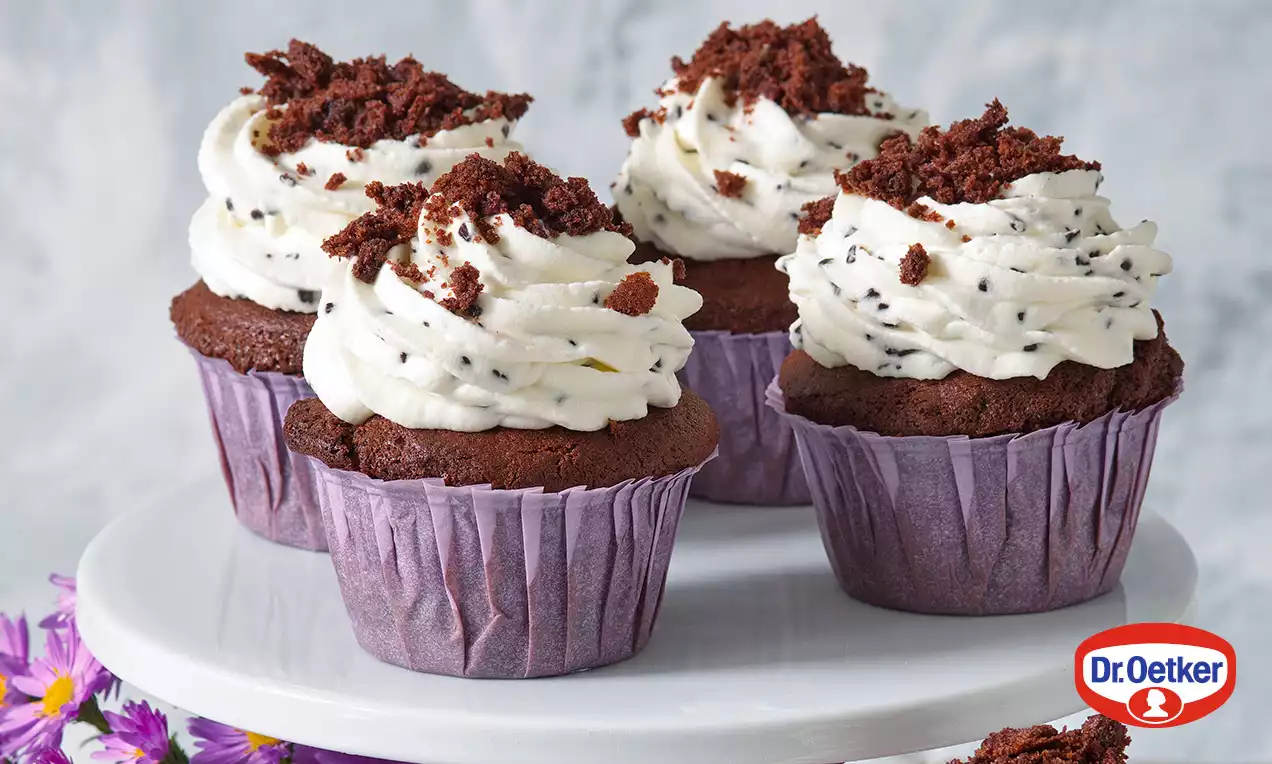 Čokoládové cupcakes s nadýchaným bielym krémom posypané tmavými čokoládovými drobkami na bielom podnose, ozdobené fialovými kvetmi