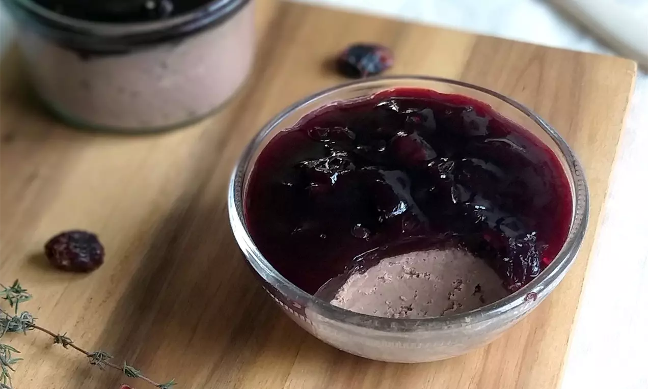 Krémová krémovohnedá krůtí paštika v sklenenej miske, pokrytá lesklým tmavočerveným brusinkovým želé na drevenom podnose s vetvičkou tymianu