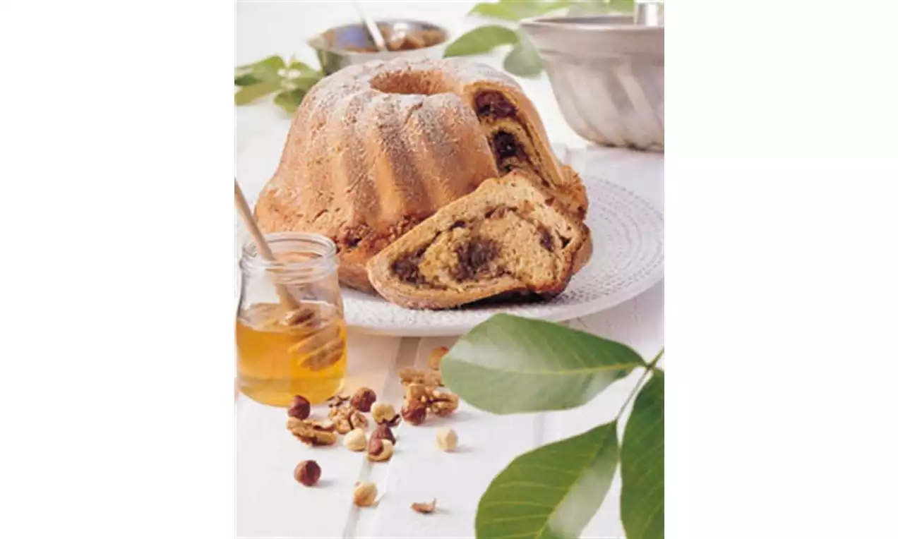 Babka s jemne hnedou kôrkou, vrstveným orechovým vírom, na bielom tanieri, s medom, lieskovcami a zelenými listami na stole