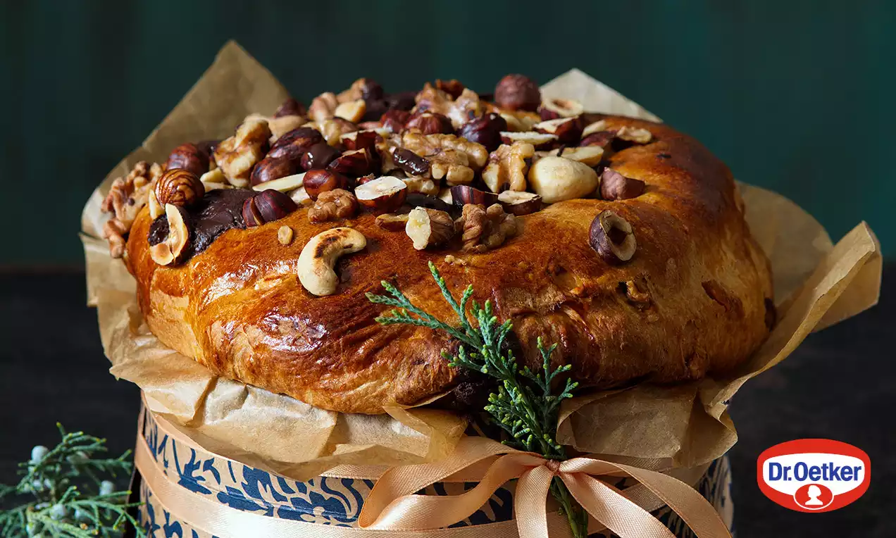 Zlatohnedý panettone s lesklým povrchom, posypaný celými orechmi, nežne uložený na jemnom papieri zdobenom modrou stuhou a vetvičkou zelene