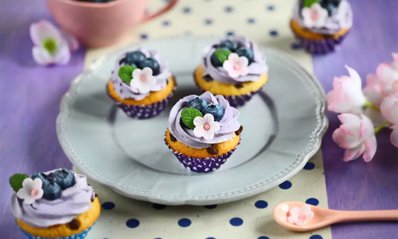 Vanilkové mini muffiny s čokoládovými kúskami zdobené fialovým krémom, jedlými kvietkami a čerstvými čučoriedkami na bielom tanieri