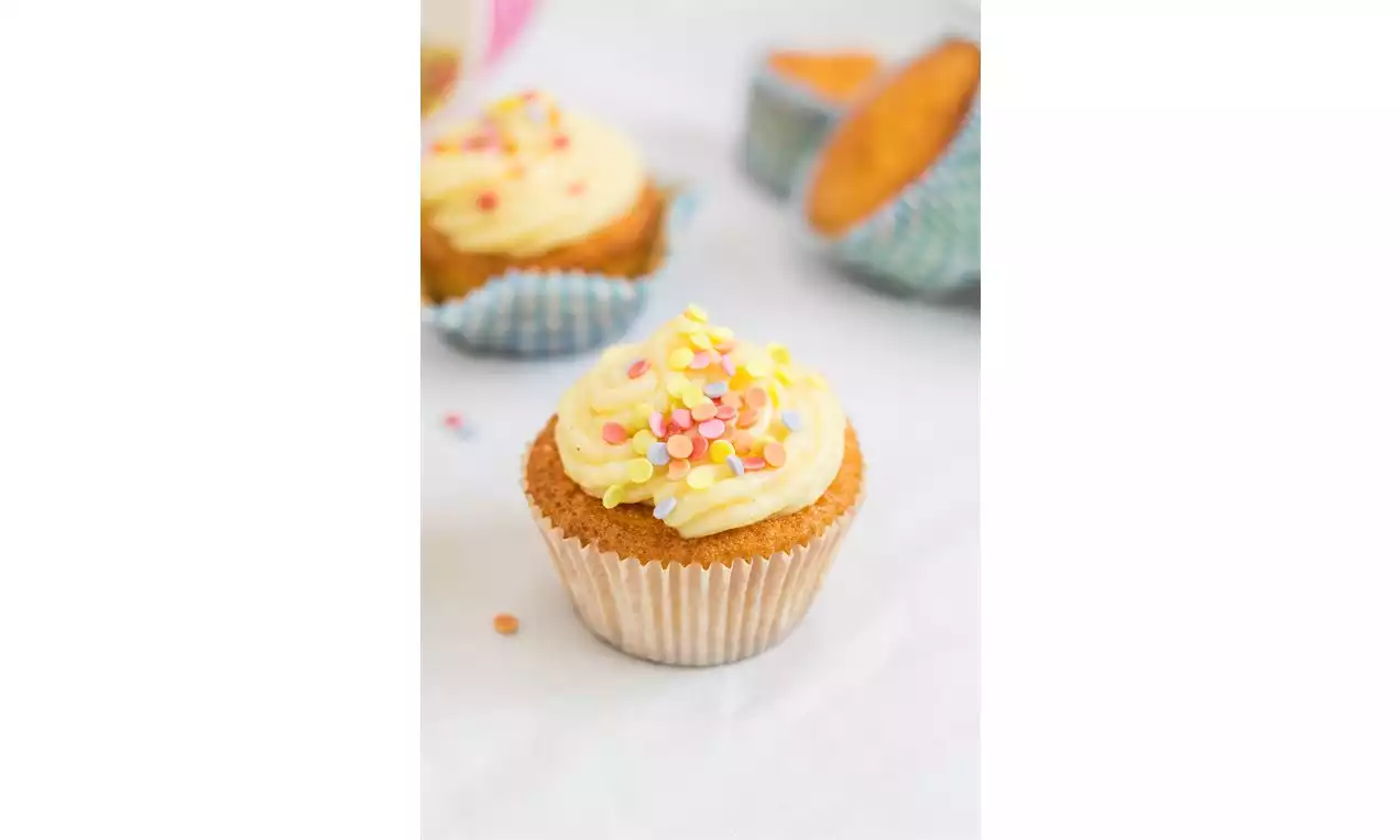Vanilkový cupcake v striebornom papierovom košíčku s jemným krémovým žltým zdobením a farebnými cukrovými posýpkami na bielom podklade