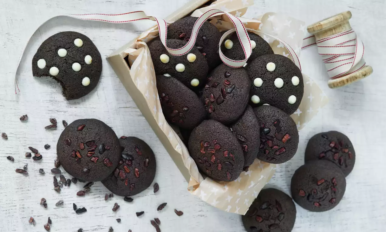 Tmavohnedé guľaté karobové cookies s hrboľatou textúrou, zdobené kakaovými kúskami a bielymi čokoládovými guľôčkami na papieri v krabičke.