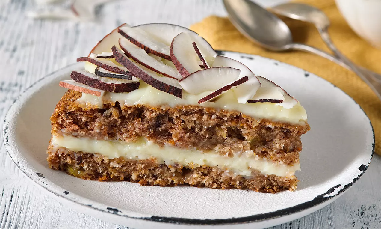 Vrstvený kúsok zlatistej mrkvovo-ananásovej torty prekladanej smotanovým krémom, ozdobený plátkami čerstvého kokosu na bielom tanieri