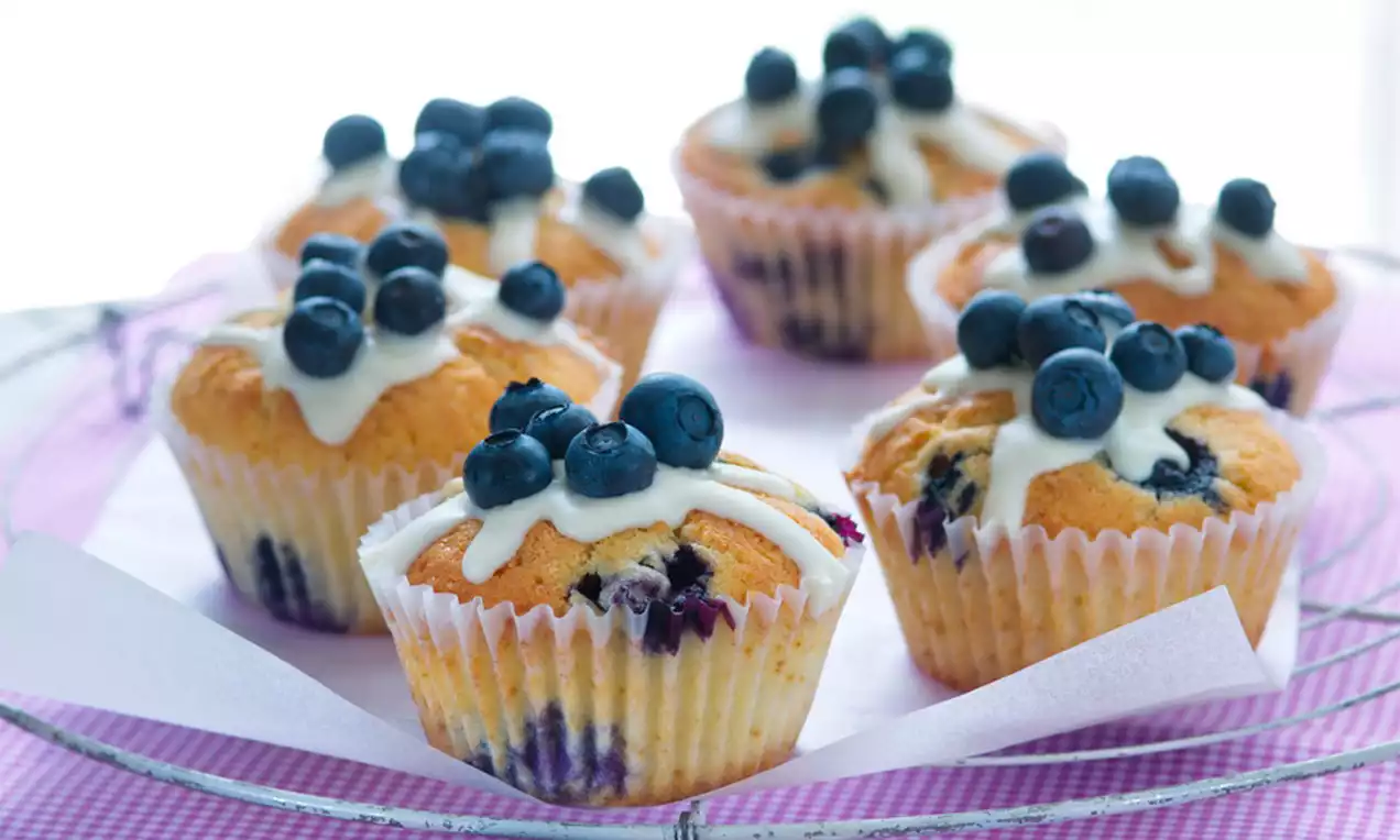 Zlatisté muffiny s nerovnomerne roztopenou bielou polevou ozdobené čerstvými tmavomodrými čučoriedkami na fialovej podložke