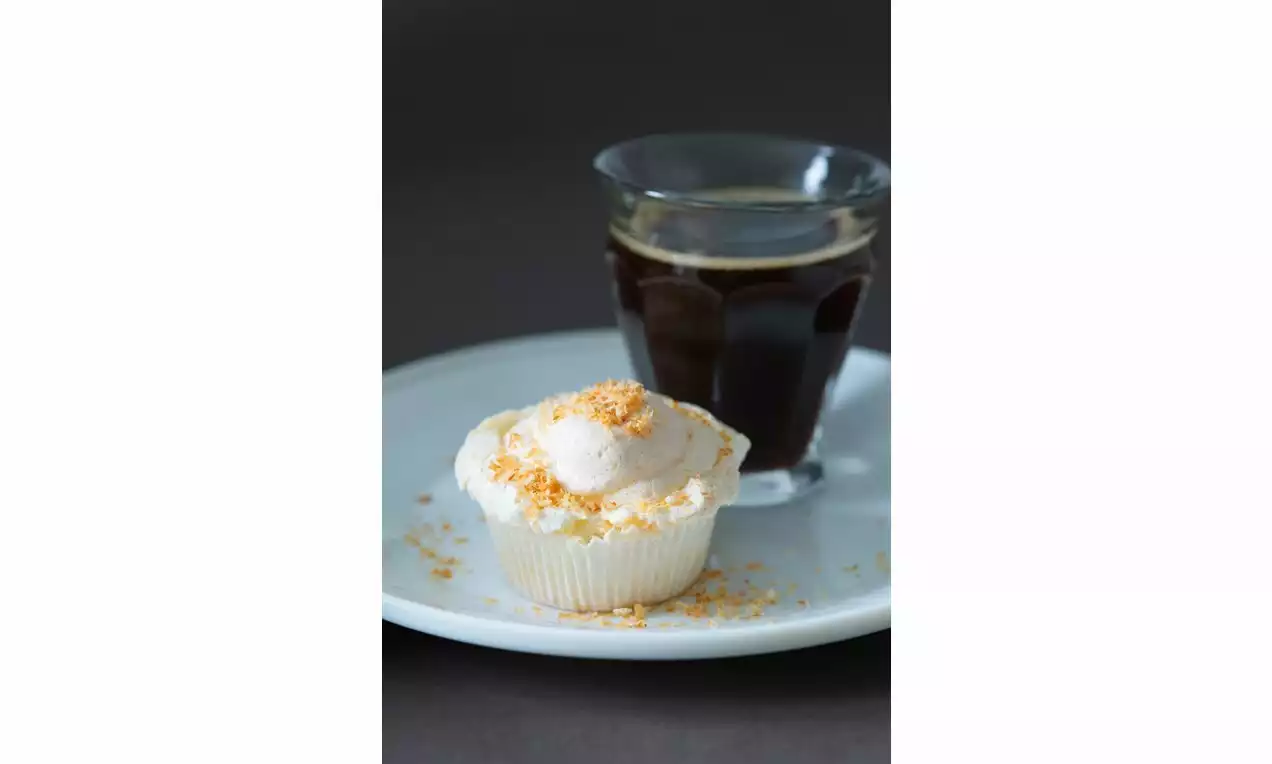 Svetlé vanilkové cupcakes ozdobené nadýchanou bielou šľahačkou s jemným posypom drobenky na bielom tanieri vedľa pohára tmavej kávy
