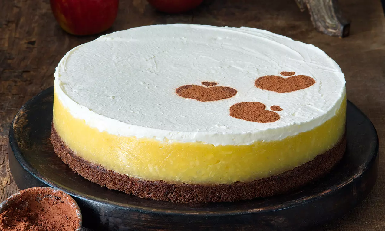 Jablkový dort s mascarpone na tmavom kakaovom korpuse s hladkou bielou vrstvou ozdobenou kakaovými tvarmi jabĺk, položený na čiernom tanieri