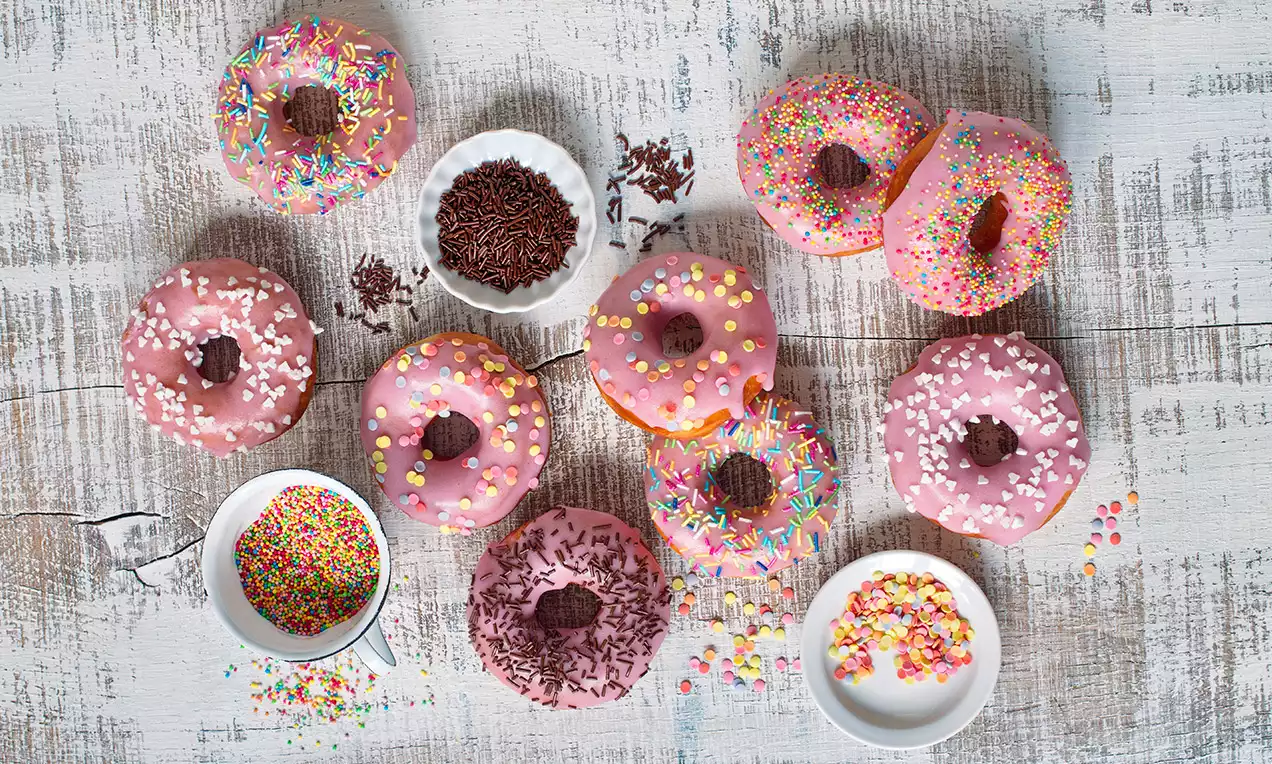 Donuty s jemne ružovou punčovou polevou, posypané farebnými cukrovými ozdobami, rozložené na svetlom drevenom podklade vedľa misiek s posýpkou