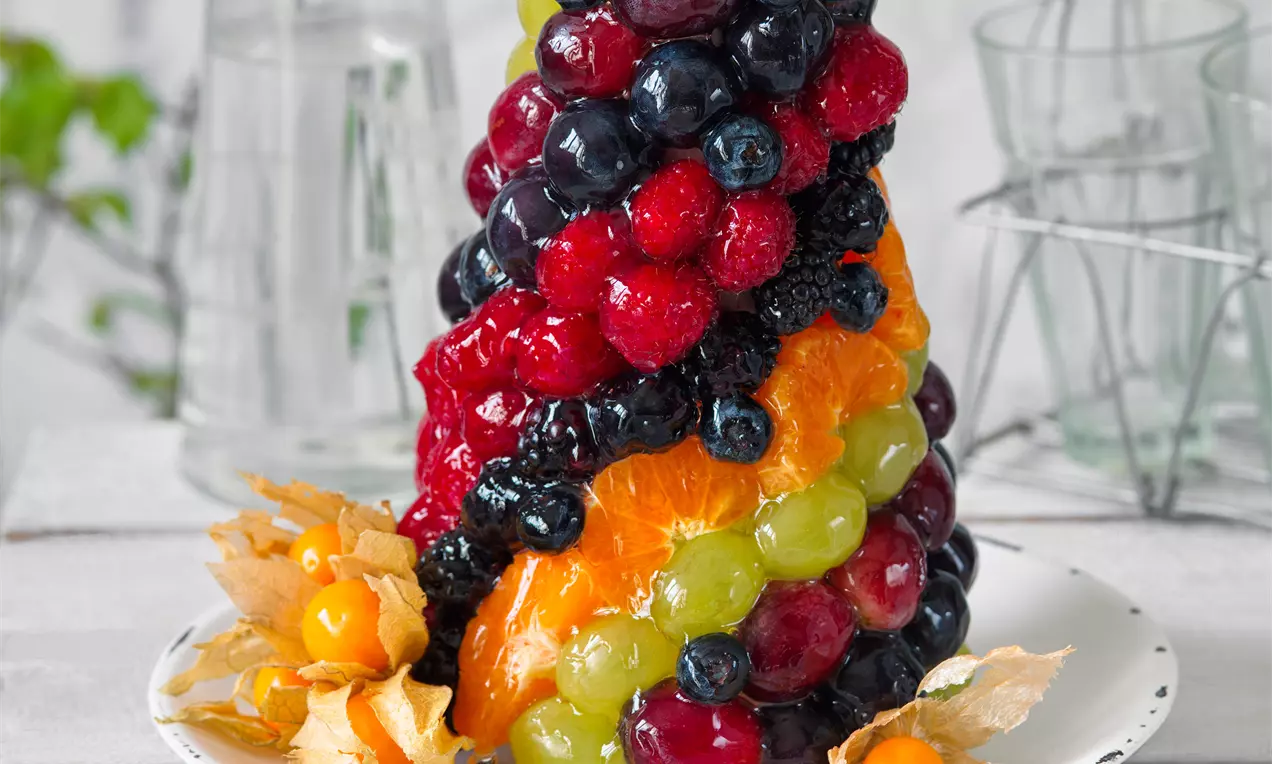 Vysoký kužeľ z lesklého ovocia v pestrých farbách, od červených jahôd po zelené hrozno, zdobený physalis, stojí na bielom tanieri