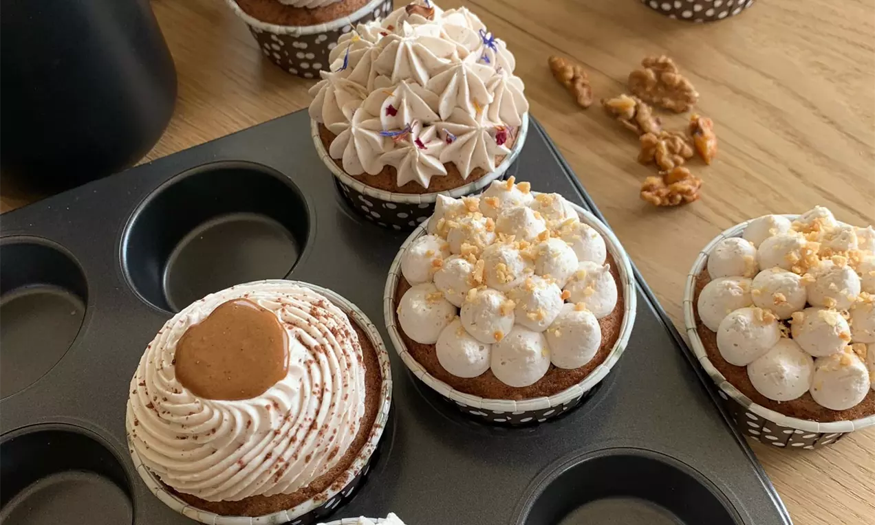 Dekorované muffiny s nadýchanými vrstvami krémov v jemných odtieňoch, zdobené orechmi, drobkami a sušienkou, v bodkovaných košíčkoch na plechu