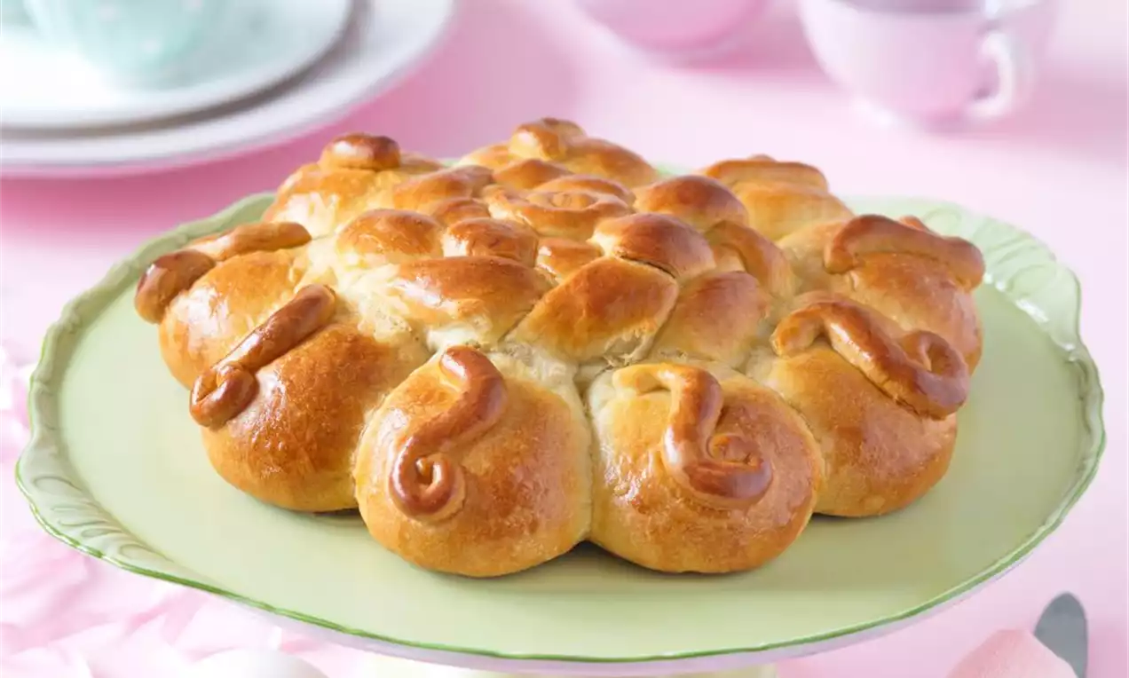 Zlatohnedé, lesklé pečivo usporiadané do guľatého tvaru na pastelovo zelenej etažéri, obklopené ružovými a bielymi veľkonočnými dekoráciami