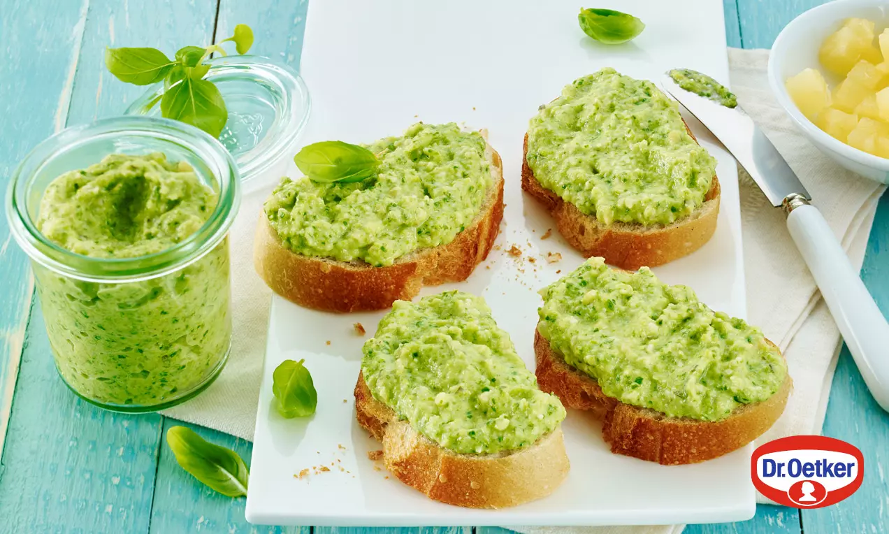 Jemné zelené pesto krémovej textúry na plátkoch opečeného chleba, podávané na bielom podnose s kúskami ananásu a čerstvými lístkami bazalky