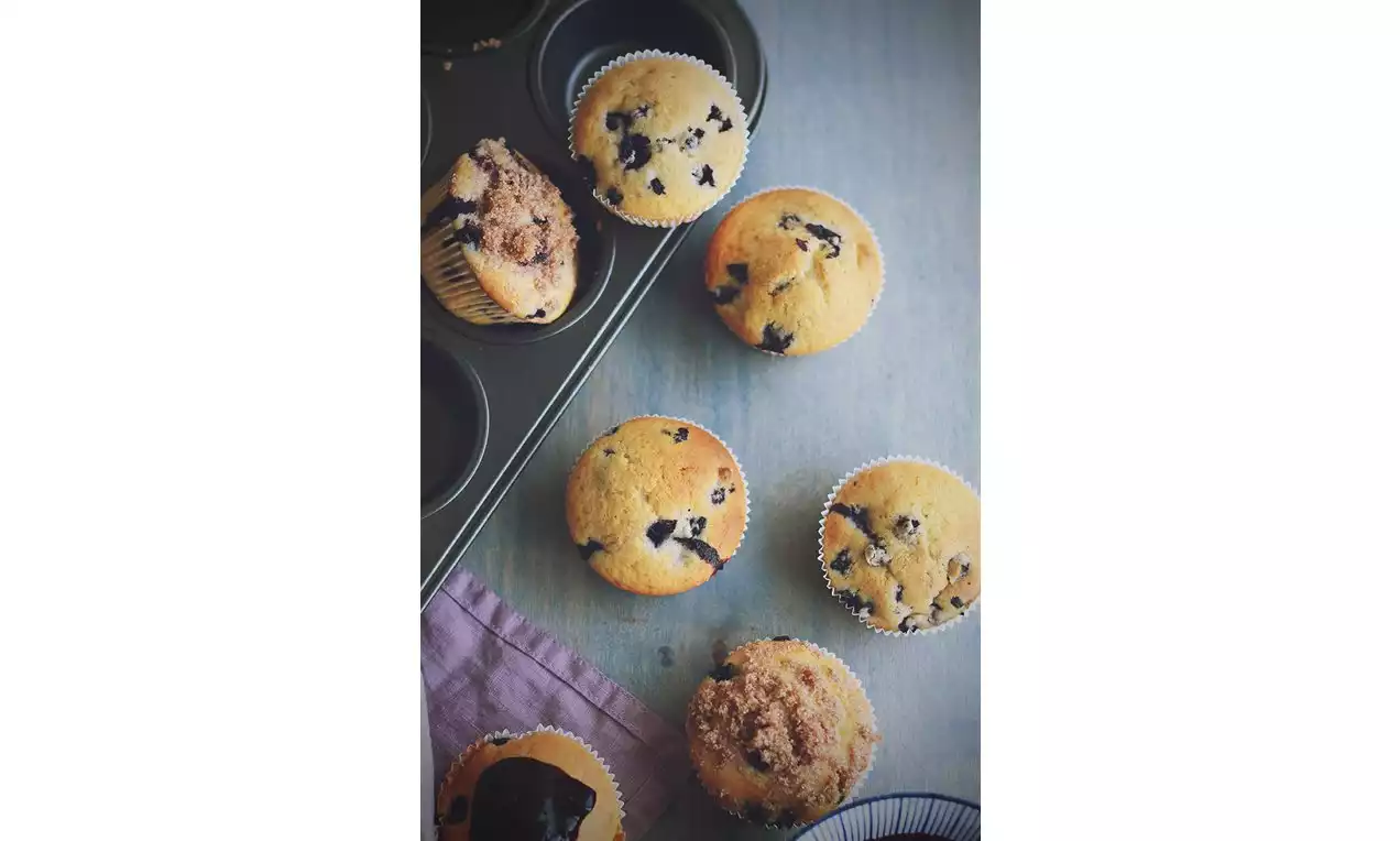 Zlatisté čučoriedkové muffiny v papierových košíčkoch, niektoré s chrumkavou drobenkou, rozložené na plechu a drevenom povrchu s fialovou utierkou