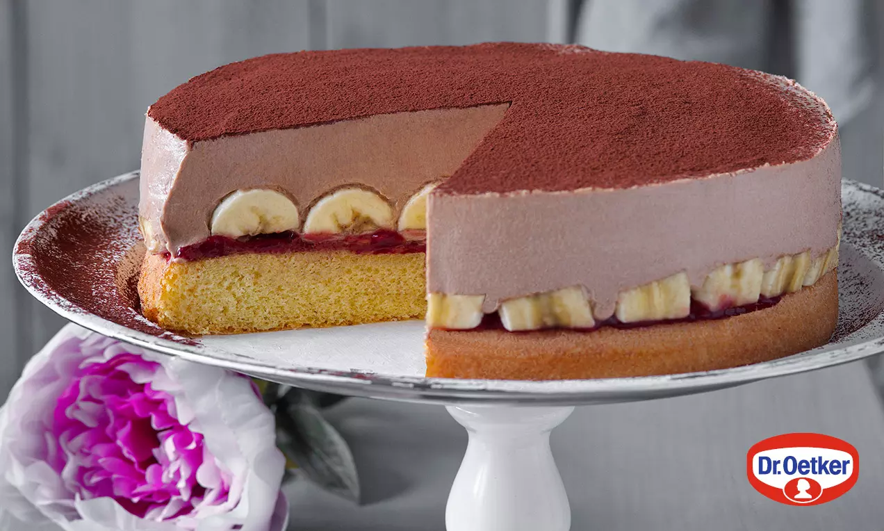 Čokoládová torta s jemnou penovou vrstvou posypanou kakaom, plátkami banánov a ovocným pyré na žltohnedom piškótovom základe