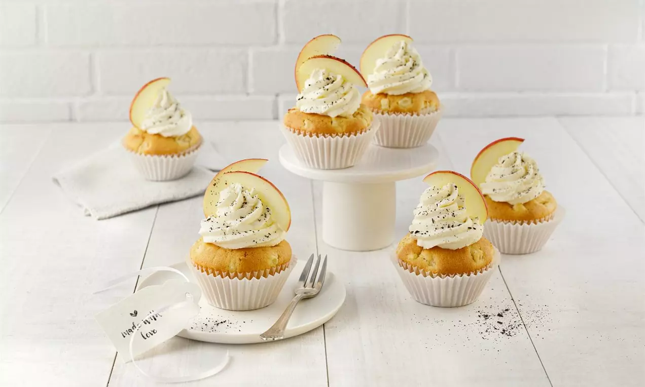 Vanilkovo-jablkové cupcakes vo svetlých košíčkoch, ozdobené nadýchanou krémovou polevou, plátkom červeného jablka a štipkou mletej škorice