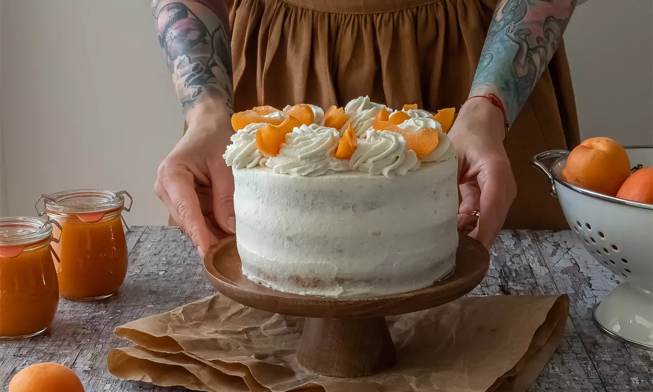 Mandľovo-marhuľová torta s jemným bielym krémom a šľahačkou, ozdobená oranžovými kúskami marhúľ na drevenom podstavci