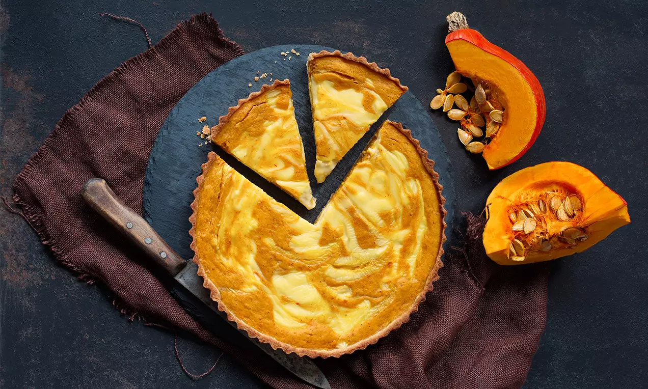 Zlatistý tekvicový cheesecake s jemne mramorovanou textúrou, nakrájaný na trojuholníky, na čiernej bridlici s ozdobou čerstvej tekvice